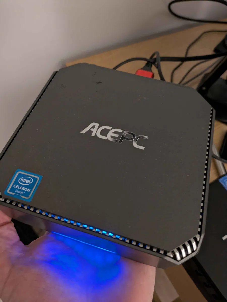 tater0, an ACEPC AK2 mini PC with an Intel Celeron sticker, held in one hand, its blue light glowing along the base